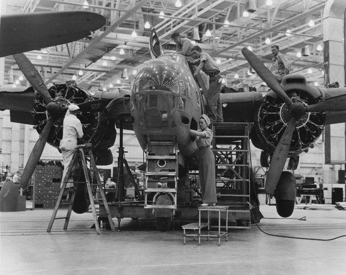 Black and white photograph of several workers working on an airplane in a plant. A few workers are on top of the plane and on the wings, and two are standing on ladders next to the plane. 