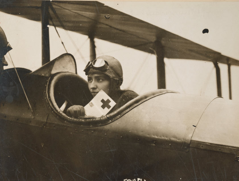 Black and white photograph of Katherine Stinson sitting in her airplane, holding a card with a cross